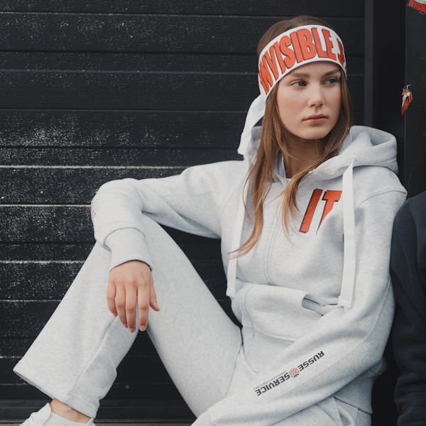 En jente sitter langs en svart vegg. Har på seg hodebånd med logo, grå henttegenser og joggebukse